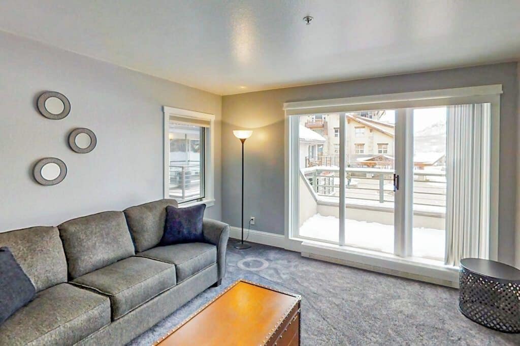 A living room with a grey sofa, a coffee table, a floor lamp, and large windows with a view of buildings.