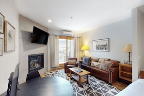 A modern hotel room with a flat-screen TV, a fireplace, a sofa, a coffee table, and a large window with curtains. - Stunning ski studio with pool, hot tub, firepit & mountain views - walk to lifts (Steamboat Springs)