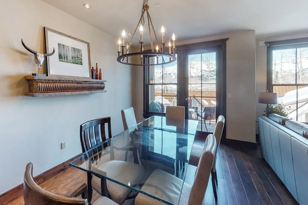 A dining area with a glass-top table, chairs, a chandelier, a mounted picture, and a window with a view.