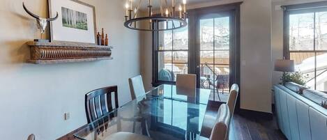 A dining area with a glass-top table, chairs, a chandelier, a mounted picture, and a window with a view.
