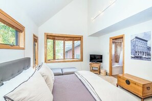 Ein Schlafzimmer mit einem Bett, einem Fernseher, einer Holzkommode und einem Fenster mit Blick auf Grünflächen.
