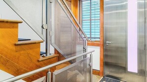 Eine Treppe mit Holzboden und Metallgeländern, eine Glastür eines Aufzugs und ein Fenster mit Jalousien.