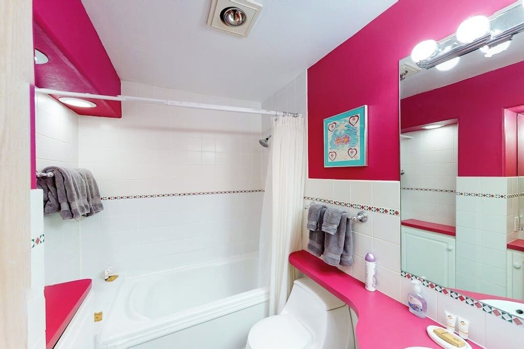 Une salle de bain avec une baignoire blanche, des toilettes blanches, un meuble-lavabo rose et un miroir avec éclairage.