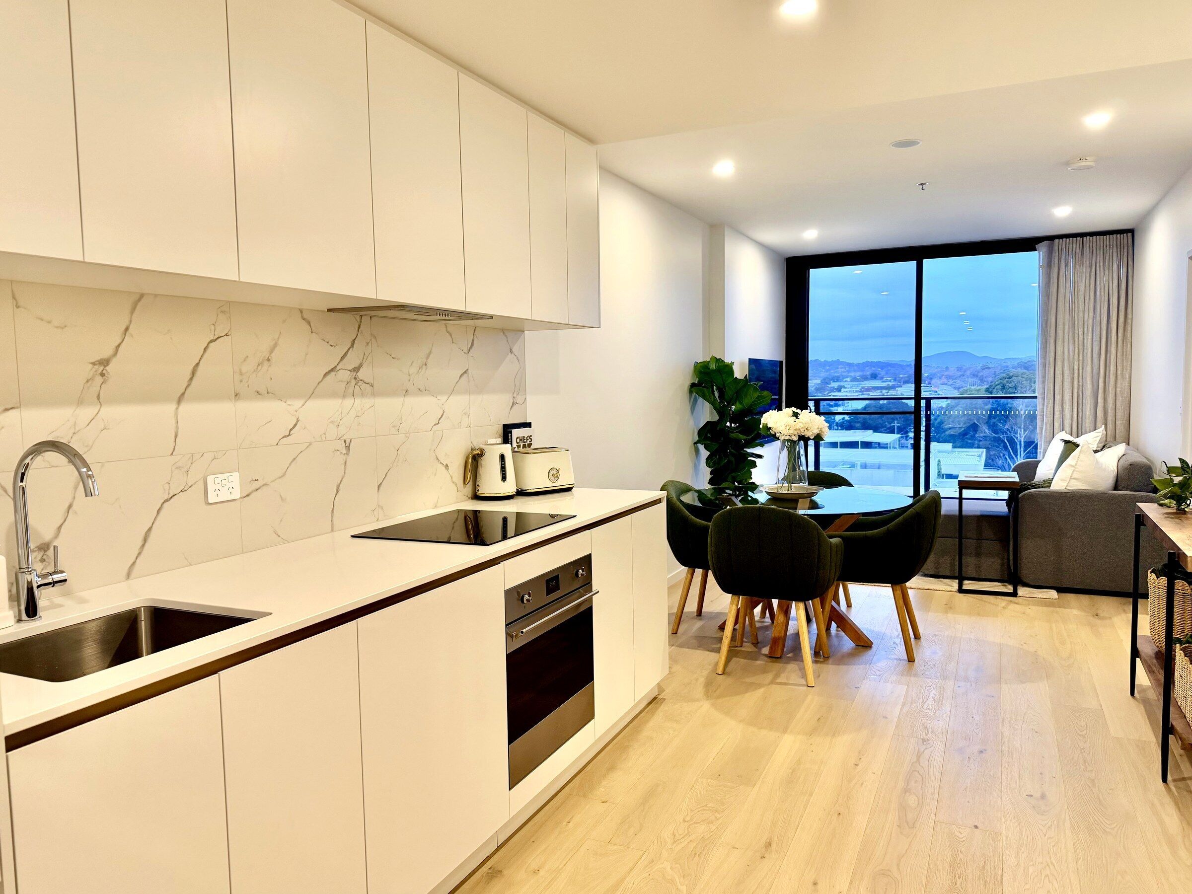A modern kitchen with a built-in oven, a dining area with a table and chairs, and a large window offering a city view.