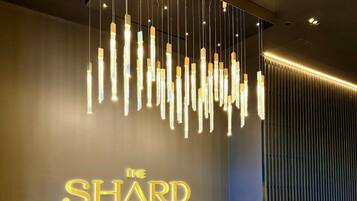 Een receptiegedeelte met een modern design, met een donkere wand waarop 'THE SHARD' in goud verlicht is, twee blauwe stoelen, een kleine ronde tafel en een unieke hanglamp.