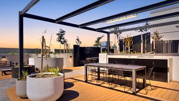 Een terras op het dak met een eettafel en stoelen, potplanten en een moderne buitenkeuken.