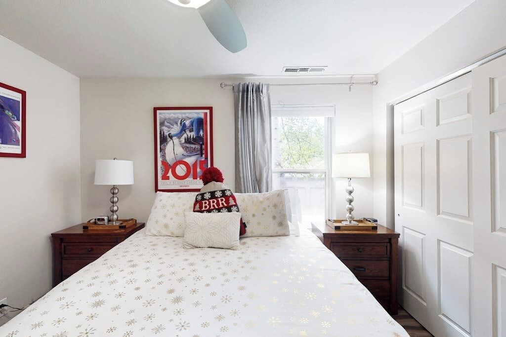 Une chambre à coucher comprenant un lit, deux tables de chevet, une fenêtre avec des rideaux, une lampe fixée au mur et un tableau encadré.