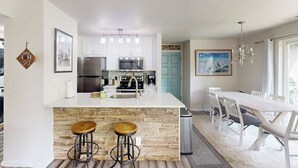 Una cocina moderna con armarios blancos, una isla central con taburetes, electrodomésticos de acero inoxidable y un comedor con una mesa y sillas blancas.