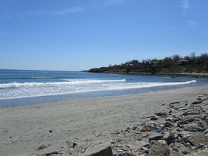 Ein Strand mit einer felsigen Uferböschung, ruhigem Meer und Häusern auf einem Hügel im Hintergrund.