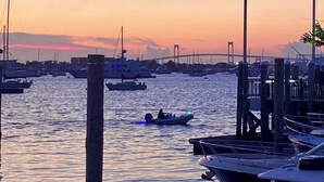 Ein Yachthafen mit vertäuten Booten, im Hintergrund eine Brücke, und ein Sonnenuntergang.