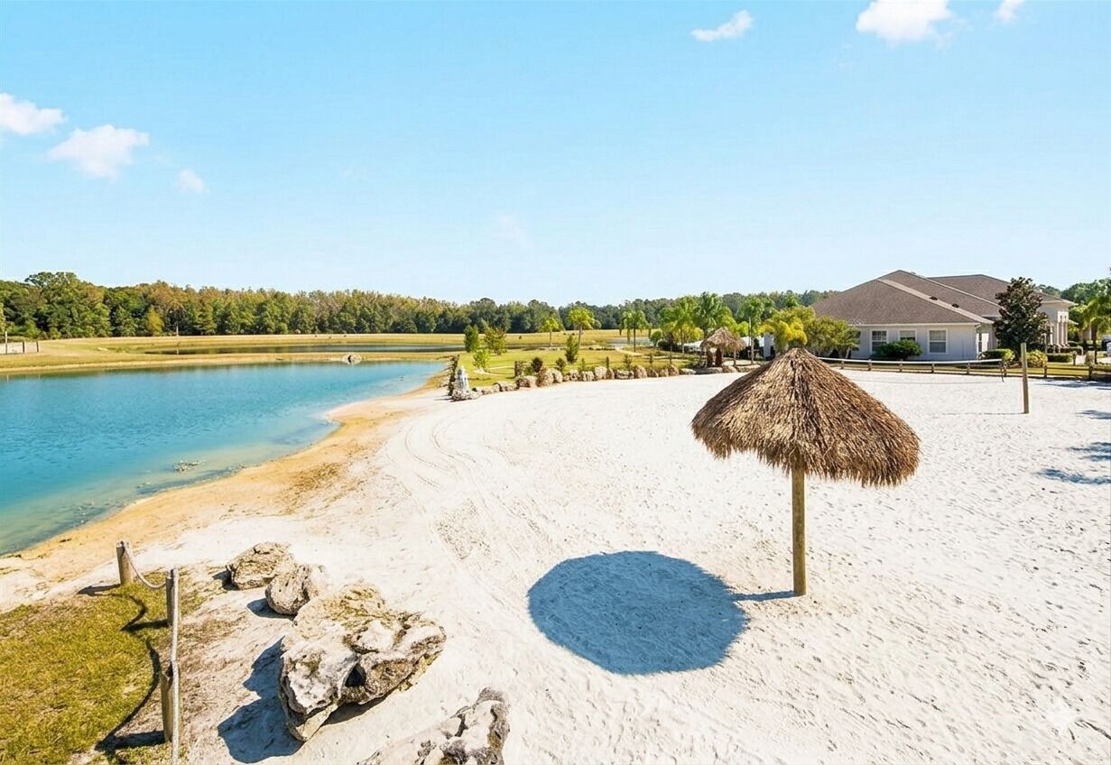 A beach with a thatched umbrella, a sandy shore, a house in the background, and a body of water.
