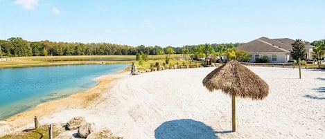 A beach with a thatched umbrella, a sandy shore, a house in the background, and a body of water.