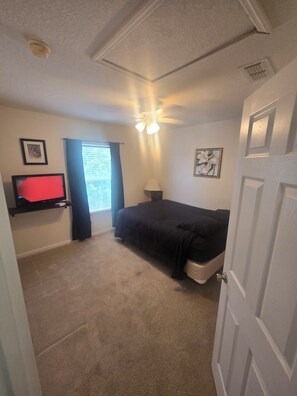 Ein Schlafzimmer mit Bett, Deckenventilator, Fenster mit Vorhängen, einem an der Wand montierten Fernseher und einer Tür.