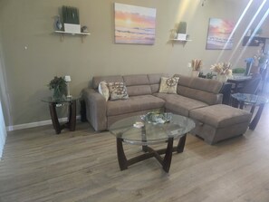 Een woonkamer met een hoekbank, een glazen salontafel en decoratieve spullen op planken.