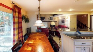 A dining area with a wooden table and chairs, a hanging light fixture, and a view of the outdoors through a window with curtains. - Creekside Condo w/ Deck: Short Drive to Skiing, Vail Village & Golf (Vail)