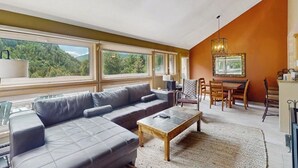 En stue med en hjørnesofa, et sofabord, et spiseområde med stole og store vinduer med udsigt over grøn vegetation.