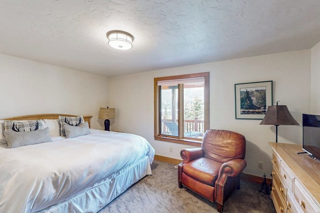 A bedroom with a bed, a leather chair, a dresser, a lamp, a TV, and a window with a view.