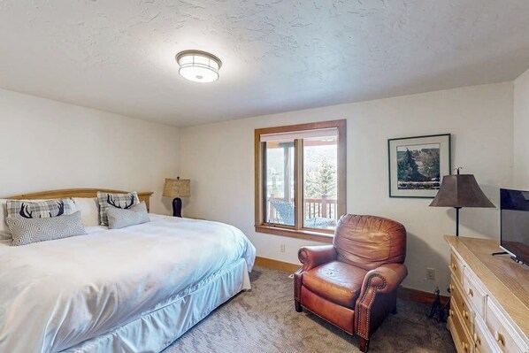 Une chambre à coucher comprenant un lit, un fauteuil en cuir, une commode, une lampe, un téléviseur et une fenêtre donnant sur un beau paysage.