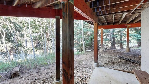 A covered outdoor area with wooden beams and columns, gravel ground, and a view of trees.
