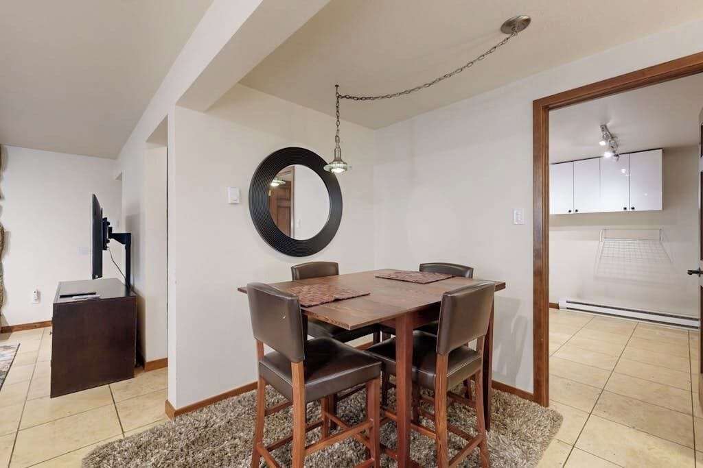 Un coin repas avec une table et des chaises en bois, un miroir rond accroché au mur et une cuisine visible à travers une porte ouverte.