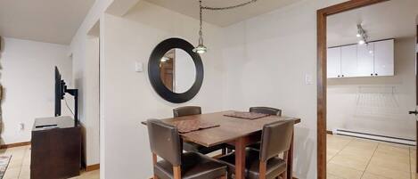 Un coin repas avec une table et des chaises en bois, un miroir rond accroché au mur et une cuisine visible à travers une porte ouverte.