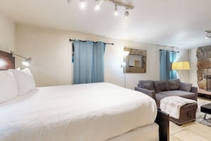 A hotel room with a large bed, a sofa, a lamp, and a stone accent wall. - Cozy Alpine Studio w/ High-End Mattresses, Hot Tub & Full Kitchen (Vail)