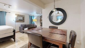 A room with a bed, a sofa, a dining table with chairs, a mirror, and a lamp.