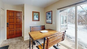 Un comedor con una mesa y sillas de madera, una puerta y un ventanal con vistas a un paisaje nevado.