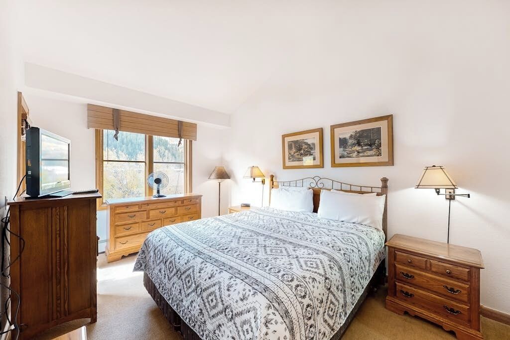 A bedroom with a bed, wooden dresser, bedside tables, and a window with blinds.
