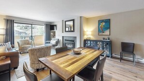 A modern living room with a wooden dining table, comfortable seating, a fireplace, and a large window. - Modern condo steps to gondola & lake w/ view & patio - in heart of everything (Avon)
