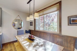 Ein Essbereich mit einem Marmortisch, Obstschale, Pendelleuchten, einem großen Fenster mit Blick auf Bäume und einem runden Spiegel an der Wand.
