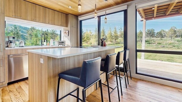 Une cuisine moderne avec des meubles en bois, un îlot central doté de tabourets de bar et de grandes fenêtres offrant une vue sur l’extérieur.