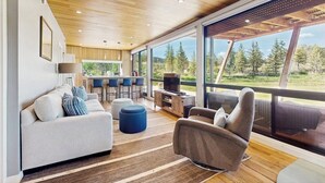 Un salon moderne comprenant un canapé, un pouf et un fauteuil, un coin cuisine et de grandes fenêtres donnant sur l’extérieur.