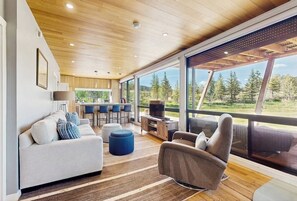 Ein modernes Wohnzimmer mit Sofa, Hocker und Sesseln, einer Küchenzeile sowie großen Fenstern mit Blick ins Freie.