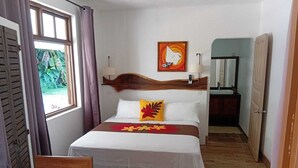 A hotel room with a bed, a wooden headboard, a window with curtains, a painting on the wall, and a bathroom visible through an open door. - Hiro'a - Fare Orotia (Raiatea)