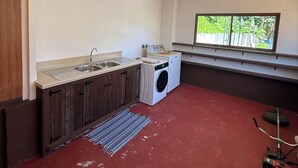 A compact kitchen with a washing machine, a window with a view, and a red floor. - Hiro'a - Fare Orotia (Raiatea)