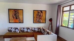 A living room with a brown sofa, white cushions, a wooden coffee table, and two framed artworks on the wall. - Hiro'a - Fare Orotia (Raiatea)