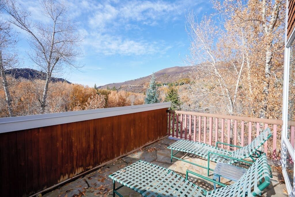 Ein Balkon mit Holzgeländer, zwei grüne Metallstühle und Blick auf eine gebirgige Landschaft.