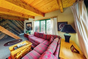 Ein gemütliches Wohnzimmer mit Holzbalkendecke, einer Sofagarnitur, einem Couchtisch aus Holz, einer Lampe und einem Fenster mit Vorhängen.