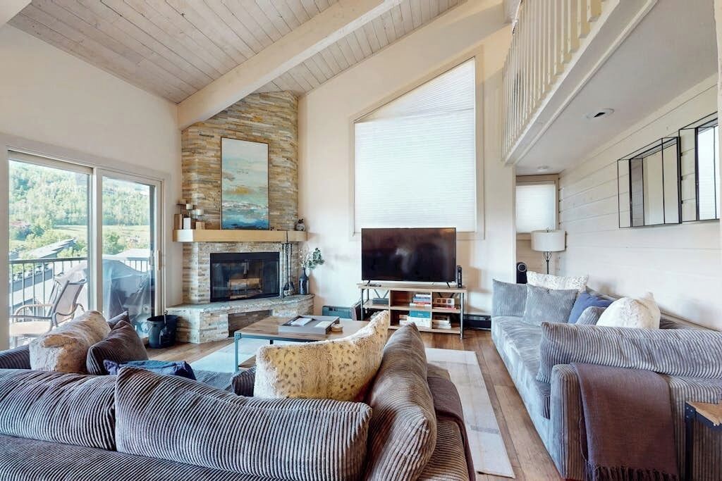 A spacious living room with a stone fireplace, a large TV, and a balcony with outdoor seating.