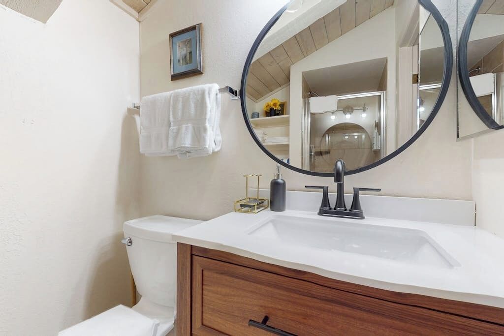 Een badkamer met een witte wastafel, een houten wastafelmeubel, een toilet en een grote spiegel met een ronde lijst.