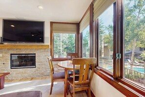 A modern living room with a stone fireplace, a flat-screen TV, and a dining area with wooden chairs. - Mountain Gem Walking Distance From Slopes w/Shared Pool & Hot Tubs, Free WiFi (Snowmass)