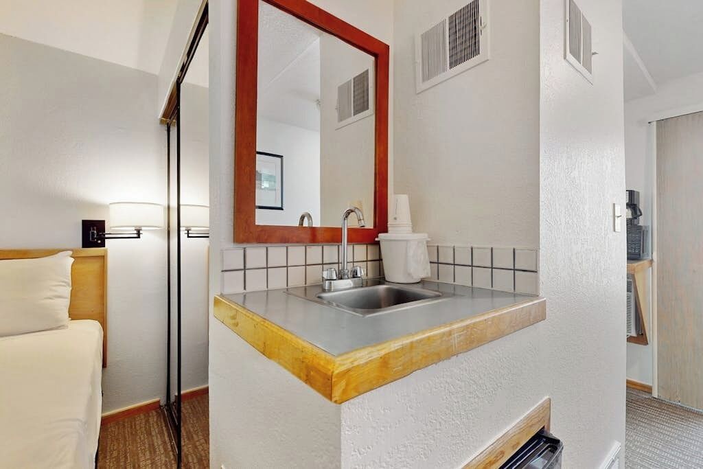 A hotel room with a wooden bed, a white sink with a red-framed mirror, and a white tiled backsplash.