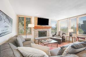 A living room with a stone fireplace, a flat-screen TV, and large windows with blinds.
