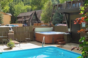 Piscina com banheira de hidromassagem, deck de madeira e vista para uma casa e floresta.