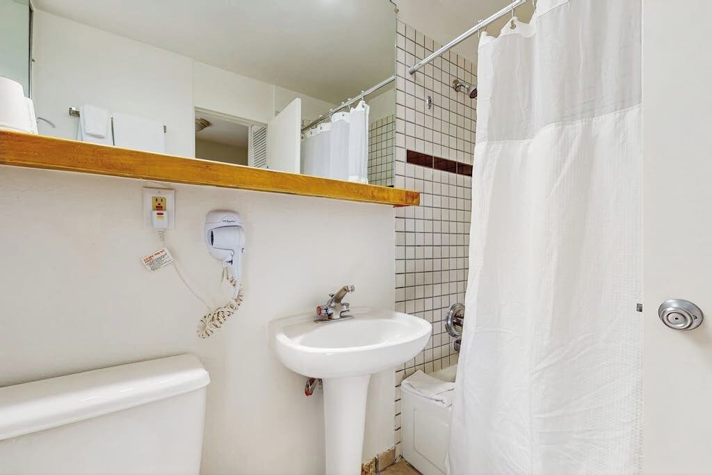 A bathroom with a white sink, a toilet, a shower curtain, and a wooden shelf above the sink.