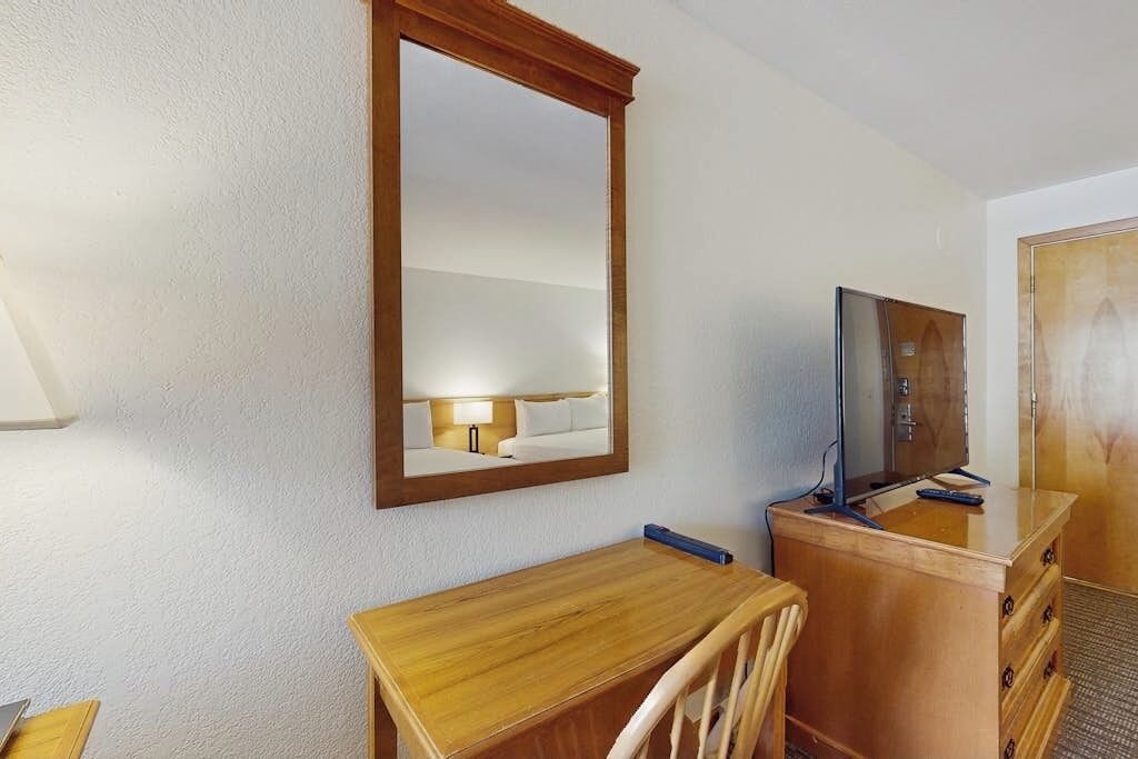 A hotel room with a wooden desk, a chair, a television on a wooden cabinet, and a large mirror on the wall.