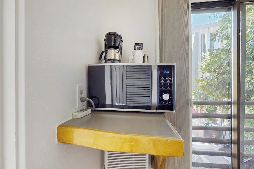 A Samsung microwave on a countertop with a coffee maker and a white appliance, a window with a view of greenery and buildings, and a yellow shelf.
