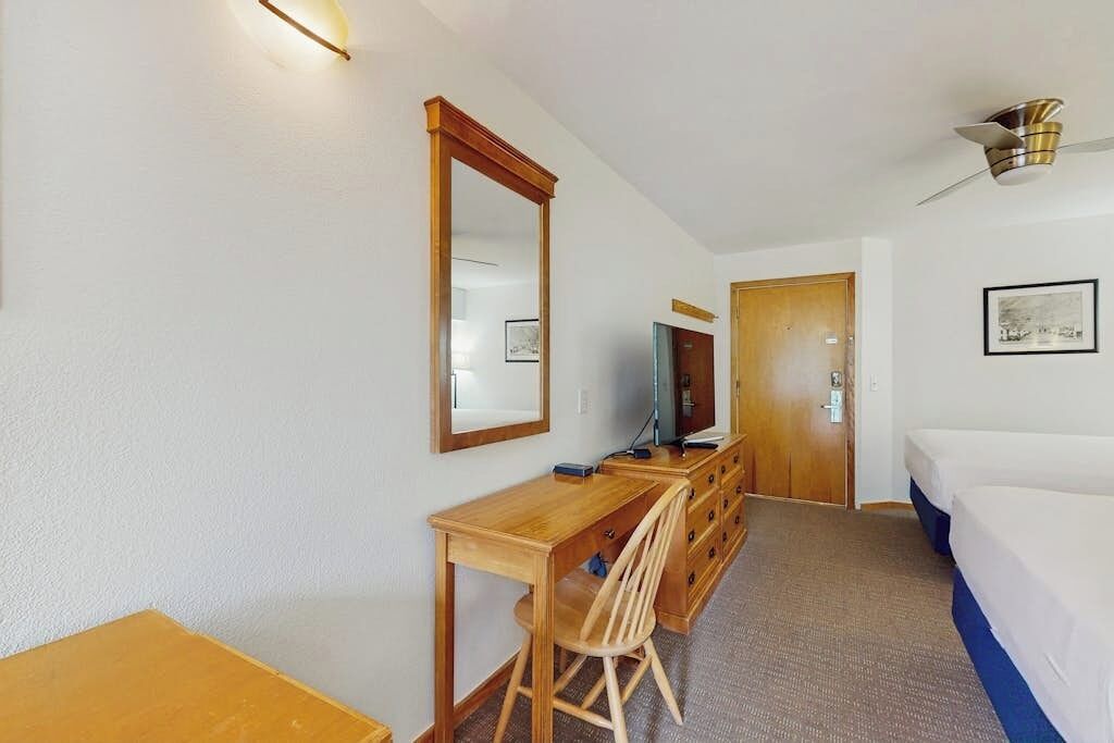 A hotel room with a wooden desk, a chair, a bed, a mirror, a television, and a door.