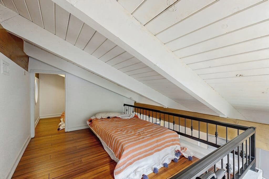 Un dormitorio con piso de madera, una cama con manta a rayas, barandal negro y techo inclinado.